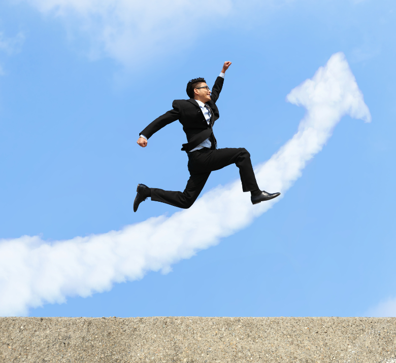 Ein Mann im Geschäftskleidung, der energisch in die Luft springt und dabei eine Siegerpose einnimmt, gegen einen klaren blauen Himmel mit einer Wolke im Hintergrund, die einem Pfeil nach oben ähnelt – ein Symbol für Erfolg und Aufstieg.